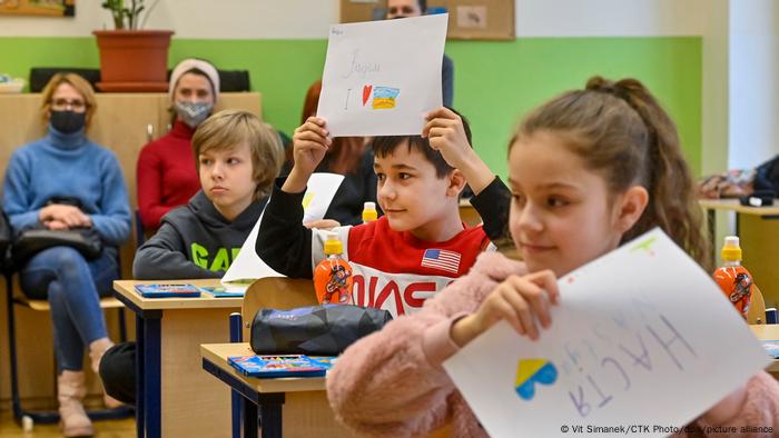 Tschechische Kinder in einer Schule in Prag halten selbst gemalte Bilder hoch. Im Hintergrund sieht man zwei Erwachsene, die das Geschehen beobachten.