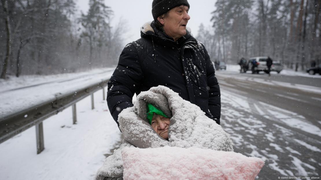 Foto mostra uma idosa muito encasacada sendo empurrada em uma cadeira de rodas por um homem. Há muita neve. 