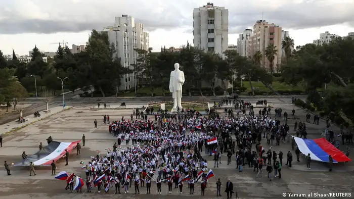 People gather near a statue of former president Hafez al-Assad, while holding Syrian and Russian flags in solidarity with Russia