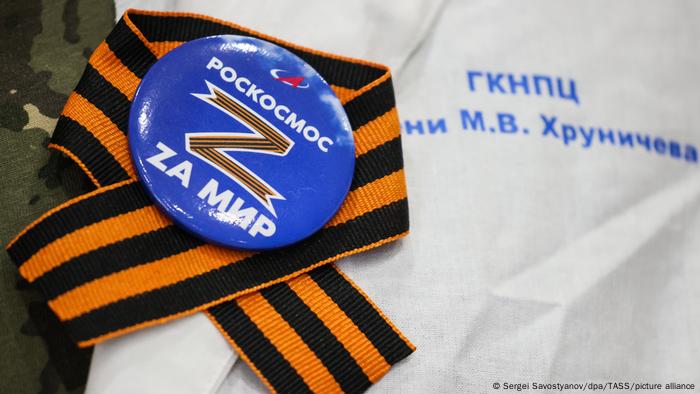 Orange and black St George ribbon with a blue bade on it that has a larf ge Z and words in Russian 