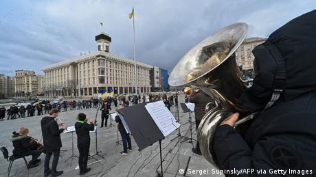 Musicians playing instruments on a alrge square, a few spectators, large building in background 