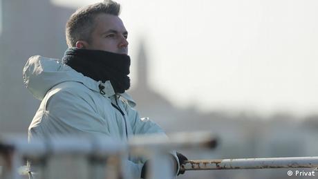 Author Sasha Filipenko wears a scarf and olive jacket, hair cropped, arm on rail