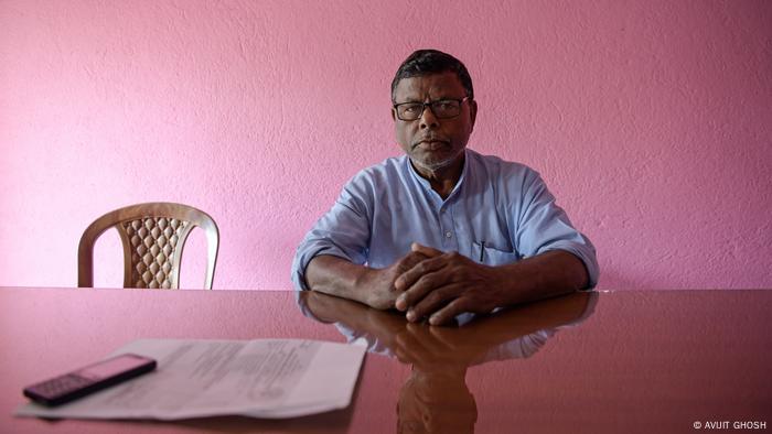 A man sitting at a desk. The wall behind him is pink 