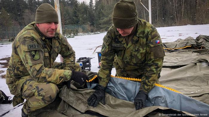 Two men in camouflage suits in the process of unfolding a tarp 