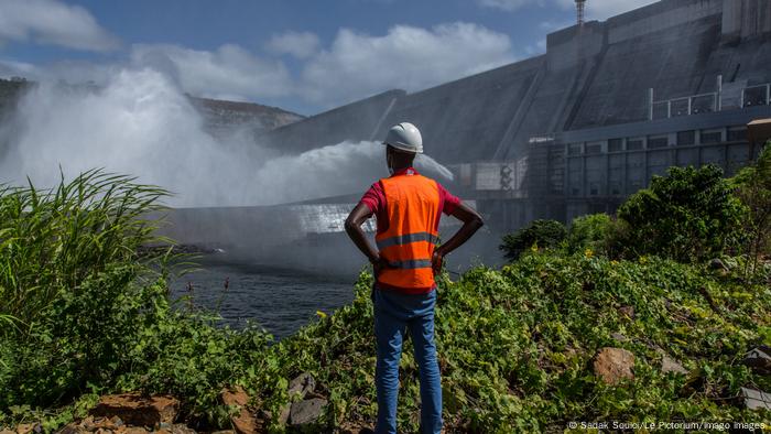 Un hombre con ropa de obra mira las nubes de vapor de agua provocadas por la caída de agua al pie de la presa de Souapiti, en Guinea.