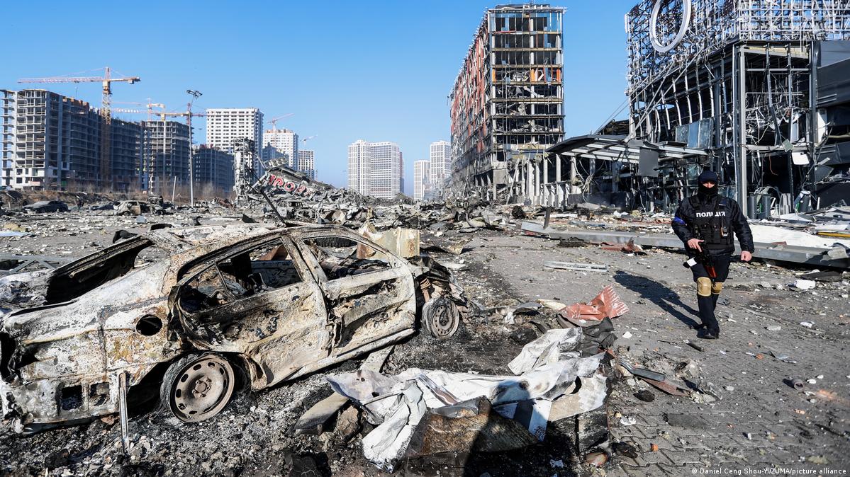 A imagem mostra um carro e uma série de prédios destruídos em decorrência de ataques russos. O céu azul ao fundo contrasta com as cores acinzentadas dos escombros. À direita, um policial ucraniano, que veste um uniforme preto, passa ao lado do veículo.