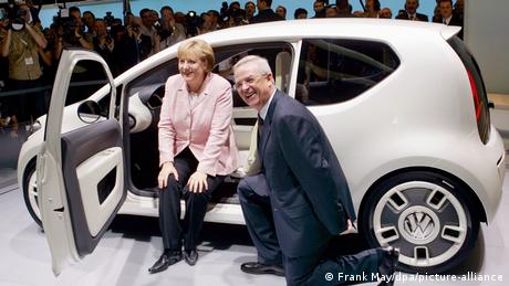 Merkel sitzt in einem VW UP und Winterkorn kniet davor auf der IAA