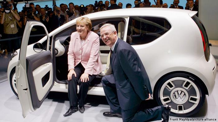 Merkel sitzt in einem VW UP und Winterkorn kniet davor auf der IAA