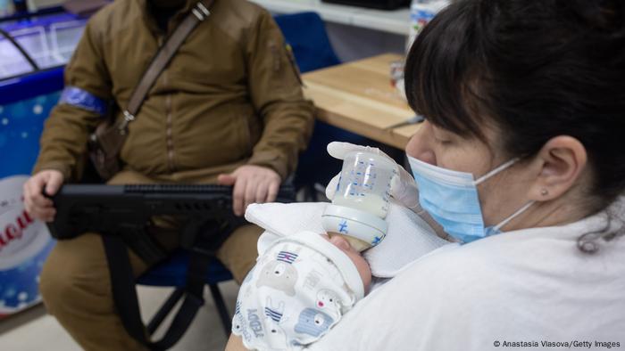 Woman with a face mask gives a baby a bottle, man with a gun visible seated in the background 