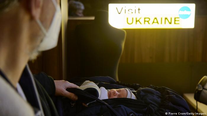 Man carrying a sleeping baby in a baby carrier, lit sign that reads 'Visit Ukraine' in the background 