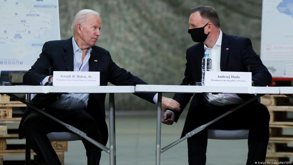 ABD Başkanı Joe Biden, Polonya'daki bir toplantıda bir masada oturuyor ve Polonya Cumhurbaşkanı Andrzej Duda'nın bileğine dokunuyor.