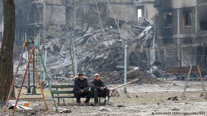 Ukrayna'nın liman kentlerinden Mariupol'da, savaşta yıkılan binaların önündeki bir bankta oturan iki kişi