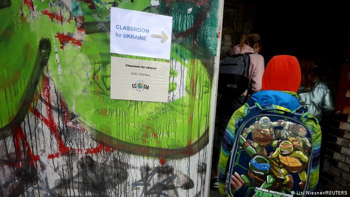Deutschland | ukrainische Schulklasse in Berlin. An einer mit Graffitis bunt besprühten Wand eines Schulgebäudes hängen zwei Zettel, auf denen Classroom for Ukraine steht. Von hinten zu sehen ist ein Kind mit Schultasche und roter Mütze, das in das Gebäude hineingeht.