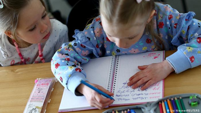 Deutschland | ukrainische Schulklasse in Berlin. Zwei aus der Ukraine geflohene Mädchen im Grundschulalter sitzen an einem Tisch in einer Schule und beugen sich über ein Heft, in das eine der beiden schreibt. 