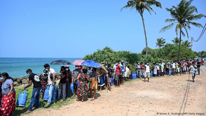 People stand in a long line in the sun between the road and the ocean