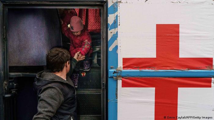 Blick in einen Bus-Eingang: Ein kleines Kind in Anorak und Mütze steigt die Treppe hinunter, gestützt von der Hand eines Erwachsenen vor ihm. Neben dem Eingang hängt ein weißes Transparent mit einem großen roten Kreuz