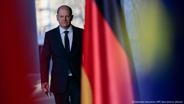  German Chancellor Olaf Scholz, paper in hand, walks as a German flag hangs in the foreground 