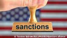 Ein symbolischer Holzstempel mit der Aufschrift sanctions, gehalten von einer Hand im Anschnitt vor einer unscharfen US-Flagge im Hintergrund. Foto mit Composing [M]. || Modellfreigabe vorhanden