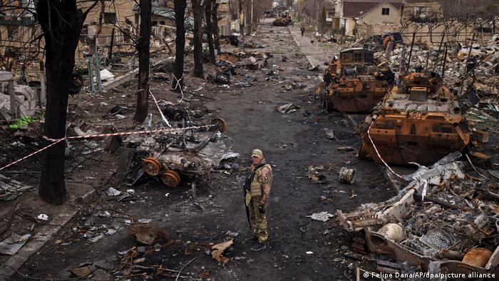 Ukraine-Krieg Butscha | Ein ukrainischer Soldat inmitten zerstörter Panzer und Ruinen, die mit Flatterband abgesperrt sind