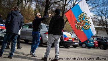 Homens na rua com bandeira russa com águia de duas cabeças, carros com bandeira ao fundo