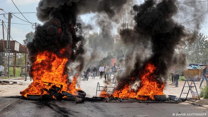 Piles of tires billow black smoke as they burn on the street with a group of Palestinian men in the background 