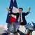 Emannuel Macron, man standing at a lectern, holds  up high both arms, flags waving in foreground