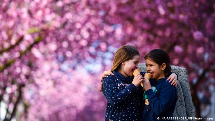Zwei Mädchen essen ein Eis und umarmen sich dabei. Im Hintergrund sind verschwommen blühende Kirchbäume zu sehen.