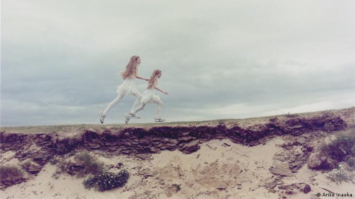 Two twin girls with long blond hair and dressed in the same white clothing run along the edge of a meadow above a sandy dropoff 