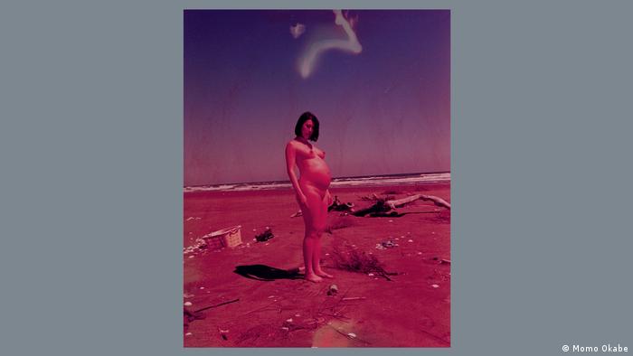A photograph dominate by red-purple tones shows a naked pregnant woman standing on a beach