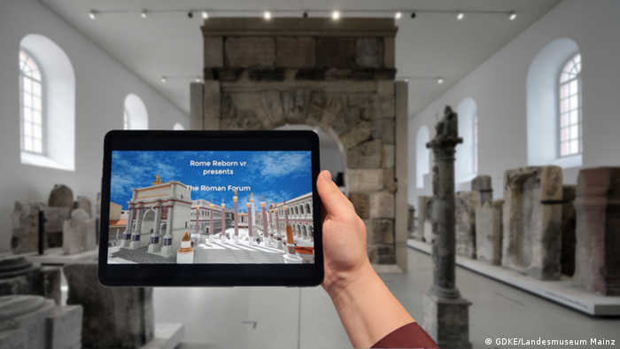 A hand holds up a tablet that shows an image of the Roman Forum.