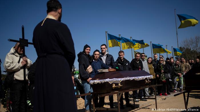 Funeral of Ukrainian soldier Anatoliy Kolesnikov presided over by an Othodox priest in black; Ukrainian flags are seen in the background