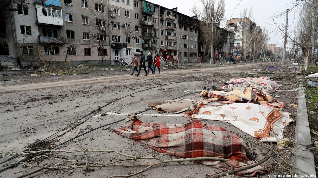Corpos tapados com cobertores em uma rua. Ao fundo, quatro pessoas caminham. Os prédios ao redor estão destruídos.