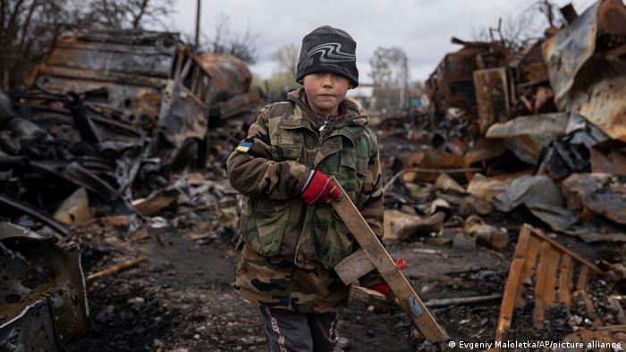 A boy wearing an army jacket holds a piece of wood like a rifle, surrounded by wrecked military vehicles