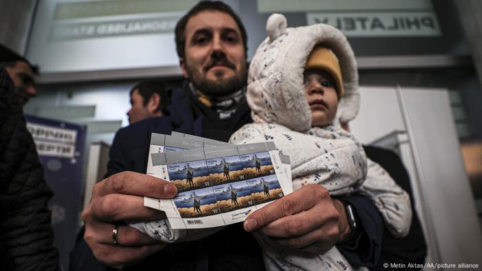 Man with child in arm shows stamps celebrating resistance of guards on Snake Island