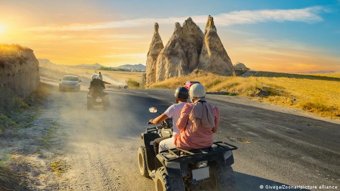 People ride quad bikes on a dusty road, in front of several rock pillars, as the sun lies low in the sky