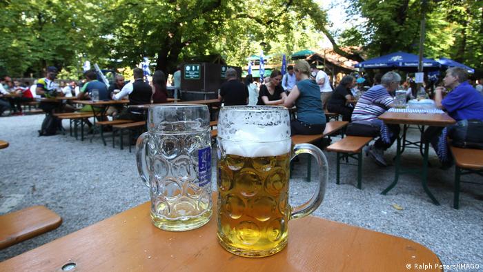 Ein leeres und volles Bierglas stehen auf einem Biertisch in einem Biergarten in München, an weiteren Tischen im Hintergrund sitzen Menschen.