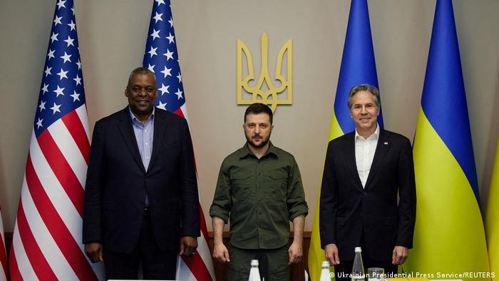 US Secretary of Defense Lloyd Austin, Ukrainian President Volodymyr Zelenskyy and US Secretary of State Antony Blinken stand in front of US and Ukrainian flags and a golden Ukrainian coat of arms