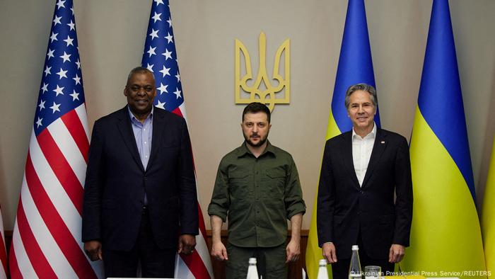 US Secretary of Defense Lloyd Austin, Ukrainian President Volodymyr Zelenskyy and US Secretary of State Antony Blinken stand in front of US and Ukrainian flags and a golden Ukrainian coat of arms