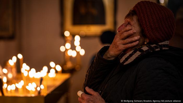 A woman wearing a knit hat covers her face as she prays in a church in Bucha, candles and icons in the background 