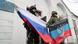 Um soldado segura uma bandeira da Rússia e outro, da ""República Popular de Donetsk"