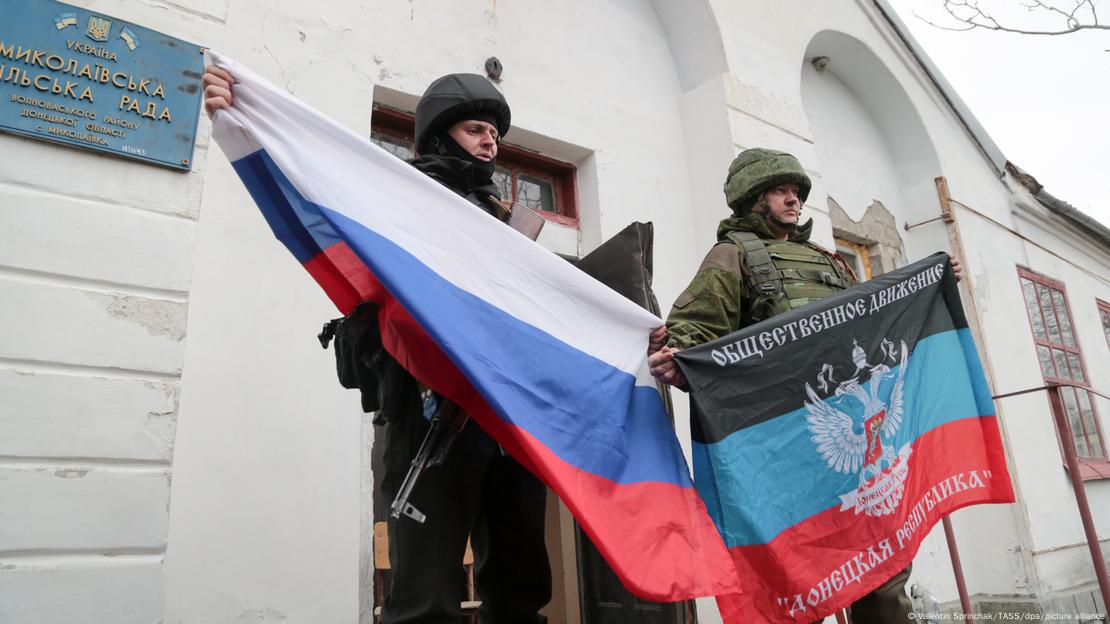 Um soldado segura uma bandeira da Rússia e outro, da ""República Popular de Donetsk"