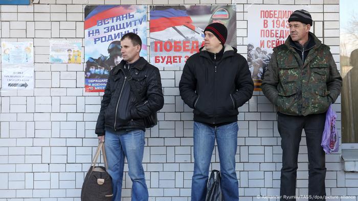 Three men in jeans and jackets wait in line with their hands in their pockets, in front of a wall with posters in Cyrillic