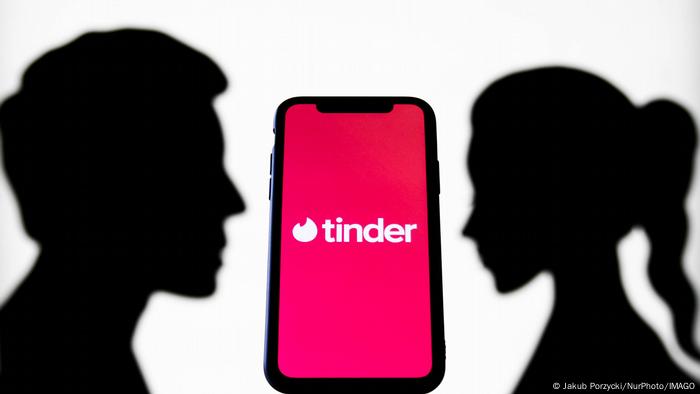 A phone showing the Tinder app on its screen stands between two dark silhouettes looking at each other