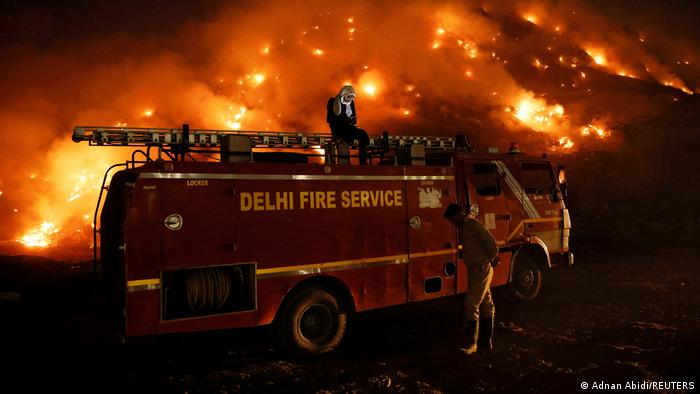 A firefighter uses his mobile phone as he sits on top of a fire truck as smoke billows from burning garbage at the Bhalswa landfill site in New Delhi, India