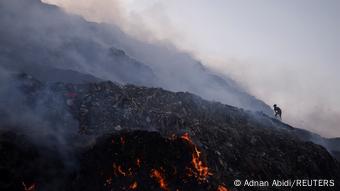 A waste collector looks for recyclable materials as smoke billows from burning garbage at the Bhalswa landfill site in New Delhi