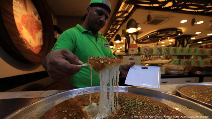 Man dripping stick over a large platter of a dark gooey dessert with crushed pistachios