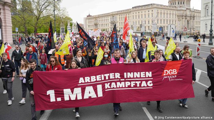 Protesters marching for workers' rights in Berlin, holding up a red banner and yellow flags