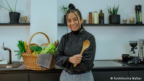 Rosa Roderigo holding a wooden spoon, standing in front of a kitchen counter, with a basket of vegetables behind her.