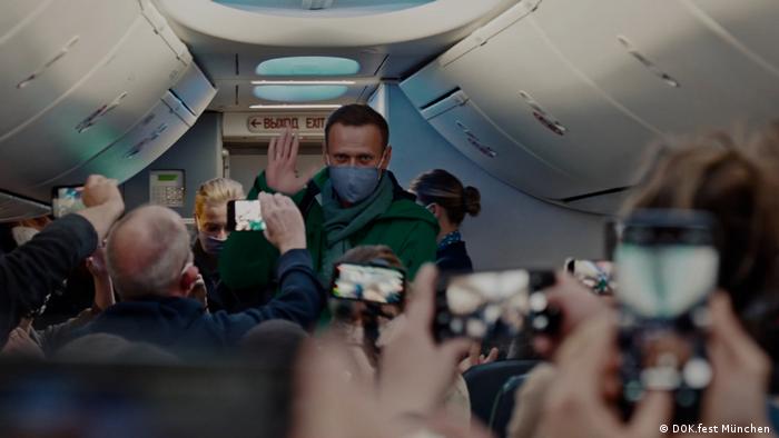 Nawalny im Gang des Flugzeugs hebt grüßend die Hand, während ihn die anderen Passagiere mit Handys filmen.