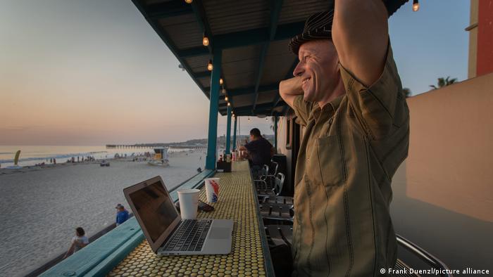 A man sits at a beach bar with his laptop, with a view of the ocean 